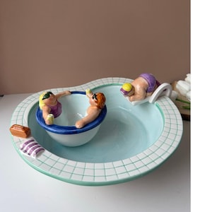 Könnte beinhalten: Eine Keramikschale im Design eines Swimmingpools mit gefliestem Rand, mit zwei Figuren in einem kleineren Becken, die gelbe Getränke halten, und einer dritten Figur auf einem Sprungbrett. Ein kleines braunes rechteckiges Objekt und ein lila gestreiftes Handtuch liegen am Rand.