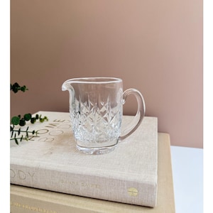 May include: A clear glass creamer with a diamond pattern. The creamer has a handle and is sitting on a book.