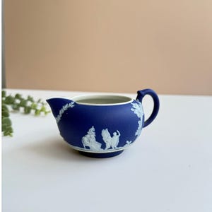 May include: A navy blue creamer with a white interior and handle. The creamer features white relief designs of classical figures and floral patterns. The creamer is a small, decorative piece, likely for serving cream or milk.
