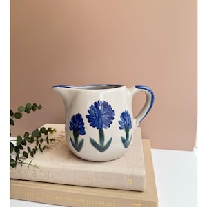 May include: Cream-colored ceramic pitcher with a blue rim and handle. The pitcher is decorated with three blue floral designs with green stems and leaves. The pitcher is sitting on top of a stack of books.