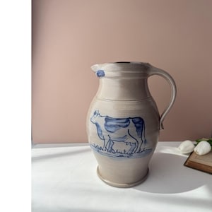 Pode incluir: Um jarro de cerâmica bege com corpo arredondado e pega curva. O jarro apresenta uma ilustração pintada à mão em azul de uma vaca num campo na parte frontal. O rebordo tem um pequeno bico e um subtil toque azul perto da pega.