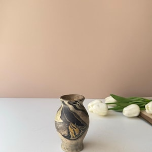 May include: A small, bulbous ceramic vase featuring a marbled pattern of black, beige, and muted gold swirls. The vase has a narrow neck and a flared rim, with a textured, matte finish throughout its rounded body and base.