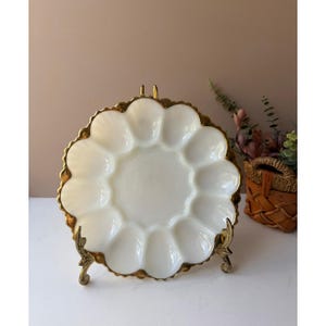 Pode incluir: Um prato de servir branco com borda ondulada e acabamento dourado, exibido em um suporte decorativo de latão. O prato tem vários compartimentos, perfeito para servir aperitivos ou lanches. A estética geral é vintage e elegante.