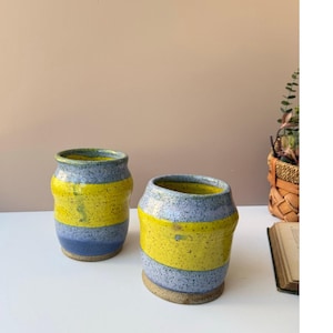 May include: Two handmade ceramic vases. The vases are cylindrical with a yellow band around the middle, blue at the bottom, and gray at the top. The interior of the vases is green. The vases are on a white surface.