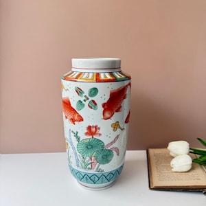 Peut inclure: Un vase cylindrique en céramique à fond blanc orné de carpes koï rouges, de nénuphars verts et de fleurs de lotus orange peints à la main. Le bord présente une bordure géométrique colorée et la base est décorée d'un motif de feuilles bleu.