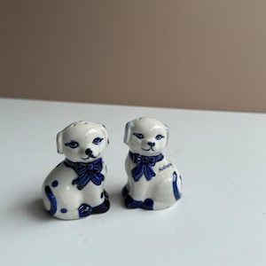 May include: Two ceramic dog figurines, white with blue accents, including spots, bows, and facial details. One figurine has the word "Solvang" on its side. These decorative salt and pepper shakers are a charming addition to any kitchen.