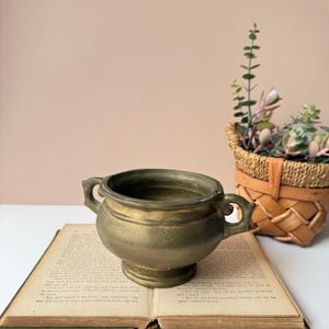 May include: A small, antique brass-colored bowl with two handles, resting on an open, aged book. The bowl has a rounded body and a pedestal base. A woven basket with greenery sits in the background.