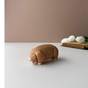 May include: A small, carved wooden pig figurine with a light brown, textured finish. The pig features a rounded body, small dark eyes, and distinct white-painted details on the snout and feet, showcasing a simple, handcrafted appearance.