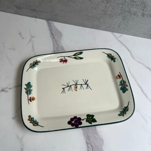 Könnte beinhalten: Eine rechteckige, weiße Servierplatte aus Keramik mit grünem Rand. Die Platte ist mit einem Blumendesign verziert, das Trauben, Kiefernäste und Beeren zeigt.
