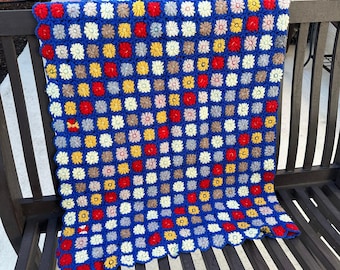 May include: A blue crocheted blanket with a scalloped edge. The blanket is made up of squares with a flower design in the centre. The flowers are in shades of white, red, yellow, and brown.