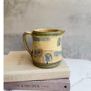 Może przedstawiać: Mały, kremowy dzbanek ceramiczny z niebieskim i zielonym wzorem. Dzbanek ma uchwyt i dzióbek. Przedstawia scenę z mostem, drzewami i budynkiem.