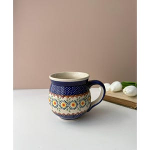 May include: A rounded ceramic mug featuring a deep blue background with a repeating pattern of white polka dots and a central band of stylized yellow and orange flowers with green leaves. The mug has a curved handle and a smooth, glossy finish.