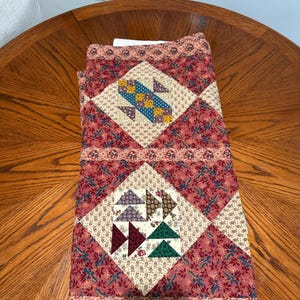 May include: A folded patchwork quilt featuring a repeating pattern of diamond-shaped blocks set against a dark red floral fabric. The quilt displays intricate geometric designs in blue, yellow, green, and purple, with a decorative border.