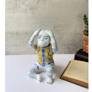 May include: A ceramic figurine of a monkey in a seated position, with its hands covering its ears. The monkey is white with blue and yellow accents, including a vest with a floral design. The figurine is on a white surface.