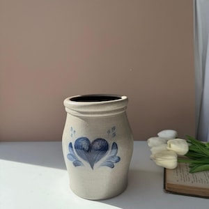 Op de afbeelding: Een beige stenen pot met een handgeschilderd blauw hartontwerp op de voorzijde. De pot heeft een afgerond lichaam, een dikke rand en een gladde, matte afwerking. Het blauwe motief bestaat uit een centraal hart omringd door bladachtige accenten.