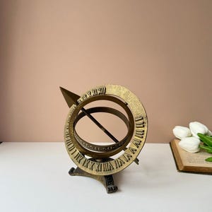 May include: A vintage brass sundial with Roman numerals etched around the outer ring. The sundial features a triangular gnomon and a circular design, resting on a decorative base. The piece is likely used for telling time based on the sun's position.