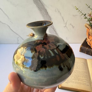 Op de afbeelding: Een handgemaakte keramische vaas met een bolvormig lichaam en een uitlopende hals. De vaas heeft een mix van blauw, bruin en zwart glazuur, waardoor een uniek, organisch patroon ontstaat. Een klein, decoratief touwtje is bij de hals bevestigd. De vaas wordt in een hand gehouden.