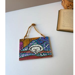 May include: Hand-painted square ceramic wall hanging featuring a whimsical cat design. The cat is white with black outlines, resting on a bed of purple flowers. The background includes shades of blue, purple, and brown, with a jute string for hanging.