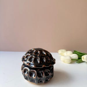 May include: A small, round, lidded ceramic jar with a dark, glossy black glaze and copper-toned highlights. The surface features a repeating, textured, dimpled pattern across both the lid and the base, creating a distinct, organic, and tactile appearance.