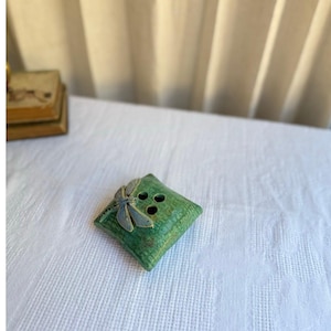 May include: A square, green ceramic flower frog featuring a textured surface and a sculpted dragonfly accent on one corner. The object has three circular holes arranged in a triangular pattern on its top surface for holding flower stems.