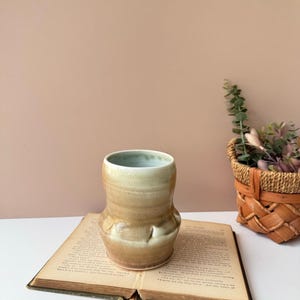May include: A ceramic tumbler with a smooth, tapered silhouette and a glossy glaze that transitions from a pale, creamy white at the rim to a warm, earthy tan at the base, featuring subtle indentations around the lower midsection.