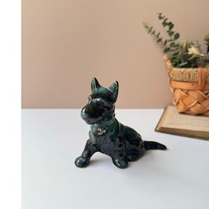 Può includere: Statuetta di cane in ceramica verde scuro lucido in posizione seduta. La scultura ha una superficie liscia e riflettente e dettagli, tra cui orecchie a punta e una coda corta. Il cane è su una superficie bianca.