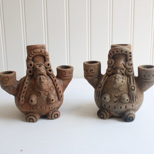 Op de afbeelding: Twee terracotta beeldjes, elk met een gestileerde vrouwelijke figuur met een gedetailleerde hoofdtooi en drie openingen. De figuren hebben armen die naar buiten uitsteken met cirkelvormige openingen en zijn versierd met decoratieve patronen.