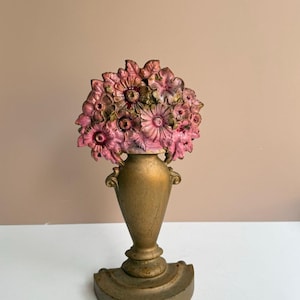 May include: A decorative bookend featuring a gold-toned vase base with a semi-circular stand. The top of the vase is adorned with a cluster of sculpted flowers painted in shades of pink, magenta, and muted green with gold highlights.
