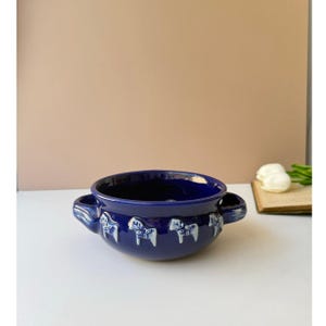 May include: A deep blue ceramic bowl featuring two side handles and a repeating pattern of white Dala horse motifs embossed around the exterior. The bowl has a smooth, glossy finish and a wide, open circular rim, showcasing a vibrant color.
