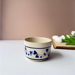 Könnte beinhalten: Eine cremefarbene Keramikschale mit einem handgemalten Muster aus kleinen blauen Beeren an dünnen grünen Ranken. Das Design wird von zwei horizontalen dunkelblauen Streifen eingerahmt, die die Außenseite umschließen, mit einem glatten Rand.