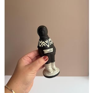 Op de afbeelding: Een zwart beeldje van een persoon met een wit patroon op het bovenlichaam en witte laarzen. Het beeldje heeft een afgeronde basis en wordt in een hand gehouden.