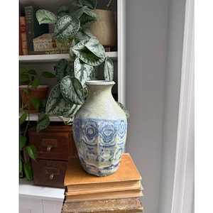 May include: A ceramic vase with a speckled light green glaze and a textured, embossed pattern around the lower body featuring deep blue circular and geometric motifs. The vase has a flared rim and a tapered neck, resting on a stack of vintage books.