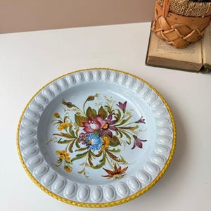 May include: A light blue ceramic plate with a yellow rim and a floral design. The plate features a raised, oval-shaped border and a central bouquet of flowers in shades of pink, blue, yellow, and green.
