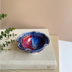 May include: A small, handcrafted ceramic bowl with a ruffled edge. The interior features a vibrant blend of red and blue hues, with a speckled, textured finish. The bowl sits on a white base and is displayed on a stack of books.