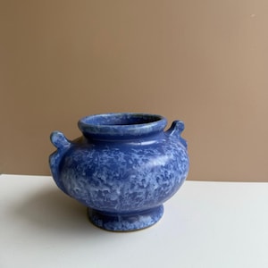 Könnte beinhalten: Eine blaue Keramikvase mit einem gesprenkelten Muster. Die Vase hat einen runden Körper, einen ausgestellten Sockel und zwei kleine Griffe an den Seiten. Der Innenraum ist ein dunklerer Blauton.