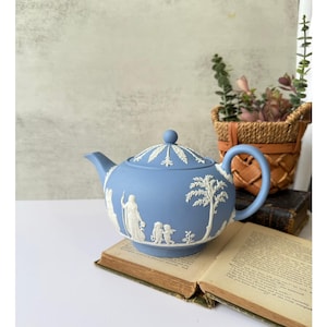 May include: A light blue ceramic teapot with white relief designs of figures and trees. The teapot has a curved spout, a rounded handle, and a domed lid with a small knob. It sits on an open book.