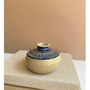 May include: A small, round ceramic vase with a speckled beige body and a dark blue rim and top. The top features a detailed, repeating pattern in dark blue. The vase is sitting on a stack of books.
