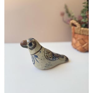 May include: A ceramic bird figurine with a brown beak and blue and brown patterned details. The bird is light beige with blue floral accents and a dark brown eye. The figurine is on a white surface.