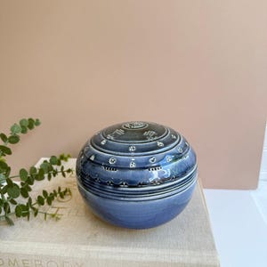 May include: A blue ceramic decorative jar with a lid, featuring intricate white patterns and designs. The jar is round and sits on top of a stack of books. The background is a neutral peach color.