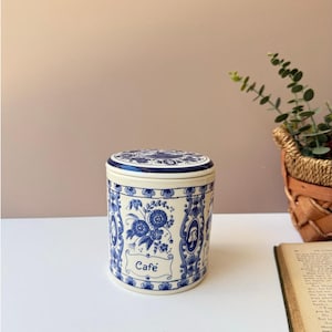 May include: A cylindrical ceramic container with a lid, featuring a blue and white floral design. The word "Café" is written in a banner on the front. The container is on a white surface.