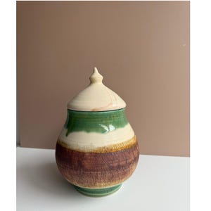 May include: A handcrafted ceramic jar with a lid. The jar features a cream-colored lid, a green band, a white band, and a textured brown base. The jar is a decorative piece.