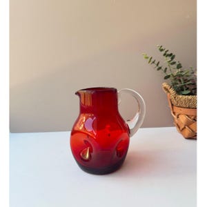 May include: A ruby red glass pitcher with a clear handle. The pitcher has a rounded body with decorative indentations. A small basket with greenery is in the background.