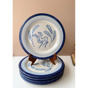 May include: A stack of ceramic plates with a blue and white design. Each plate features a blue rim, a light blue inner ring, and a central illustration of a deer and foliage in blue. The plates are stacked on a wooden stand.