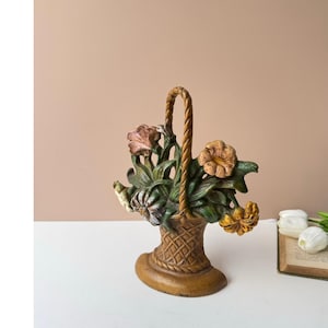 May include: A cast metal doorstop shaped like a basket filled with flowers. The basket has a woven texture and a rope-style handle. The flowers are painted in shades of pink, yellow, and white, with dark green leaves, all set on an oval base.