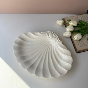 May include: A white ceramic dish shaped like a large scallop shell with a decorative embossed hinge detail at the top. The dish features deep, radiating fluted ridges that extend from the center to the scalloped edges of the shell form.