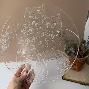 May include: Clear glass plate featuring an etched design of five cats. The cats are depicted with detailed fur and facial features, some wearing collars and a bow tie. The plate is round and appears to be vintage.