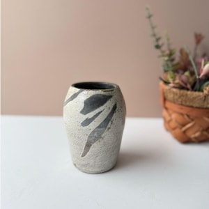 Könnte beinhalten: Eine kleine, cremefarbene Keramikvase mit dunkelgrauen, abstrakten Markierungen. Die Vase hat eine leicht strukturierte Oberfläche und ein dunkles Inneres. Ein dekorativer Artikel für die Inneneinrichtung.