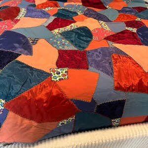 May include: A colorful patchwork quilt with a variety of fabrics in shades of red, orange, blue, and purple. The quilt features a geometric design with irregular shapes and visible stitching. The fabrics include satin, velvet, and floral prints.