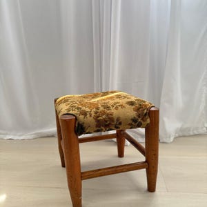 Könnte beinhalten: Ein kleiner Holzhocker mit vier zylindrischen Beinen und einer quadratischen Sitzfläche, die mit einem strukturierten Stoff mit braun-goldenem Blumenmuster bezogen ist. Der Holzrahmen hat ein mittelbraunes Finish und horizontale Streben.