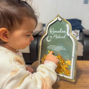 May include: A child reaches for a gold star on a Ramadan Mubarak sign. The sign is green with gold accents and features a crescent moon and star.